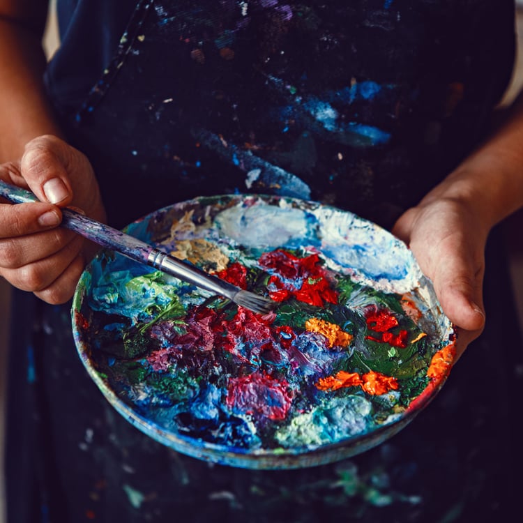 Hands of female artist holding messy dirty palette  with different paints and paintbrush in art studio. Lifestyle and hobby concept