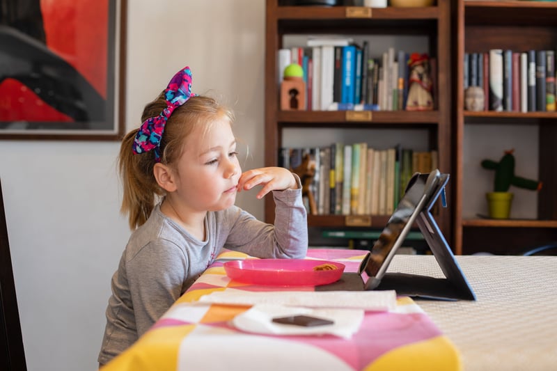 four years old blonde girl with bow diadem sitting indoor in living room, watching digital tablet on table, with library