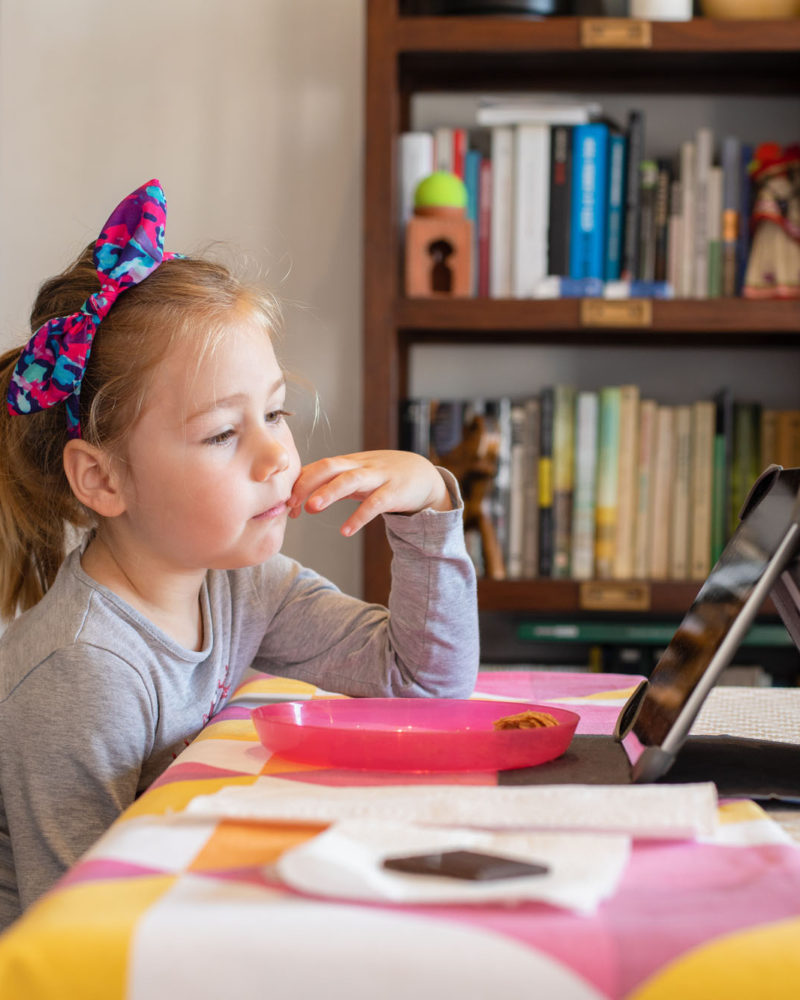 four years old blonde girl with bow diadem sitting indoor in living room, watching digital tablet on table, with library