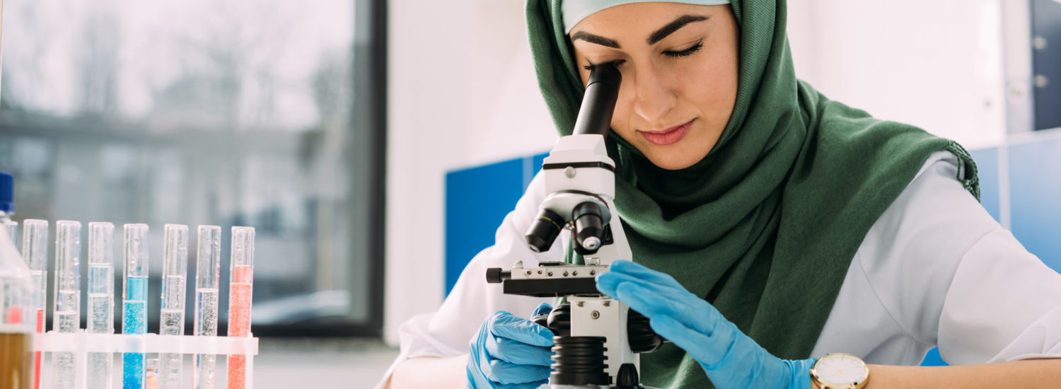 beautiful female muslim scientist looking through microscope during experiment in chemical laboratory