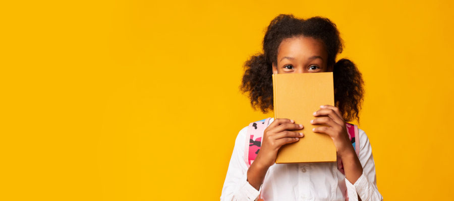 Shy African American Schoolgirl Covering Face With Book On Yellow Background In Studio. Panorama With Empty Space
